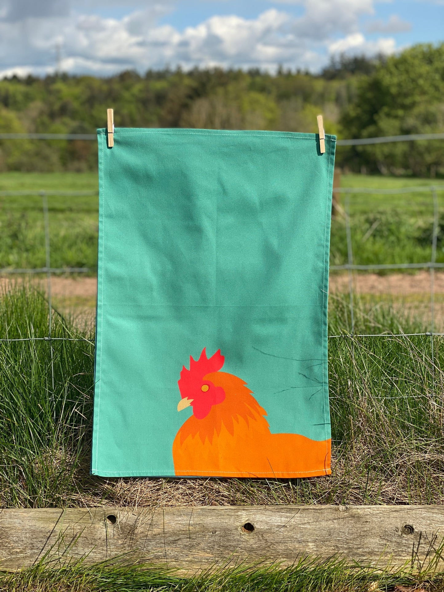 Green Chicken Tea Towel hanging by pegs on fence with countryside behind.