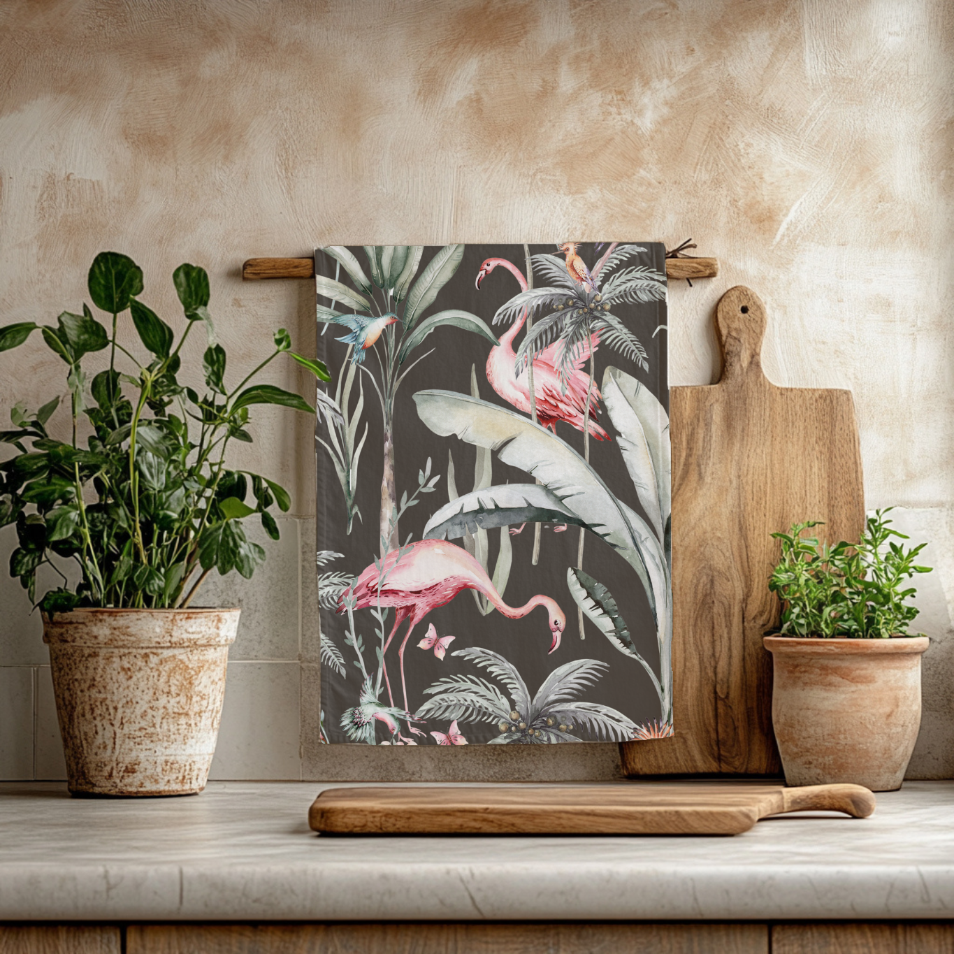 Dark Tropical Flamingo Tea Towel hanging on beige background with two potted plants and two wooden chopping boards.  