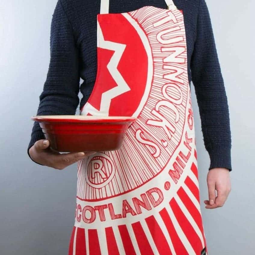 Tunnocks Tea Cake Wrapper Apron being worn by a man wearing a navy jumper and holding a casserole dish in his hand.