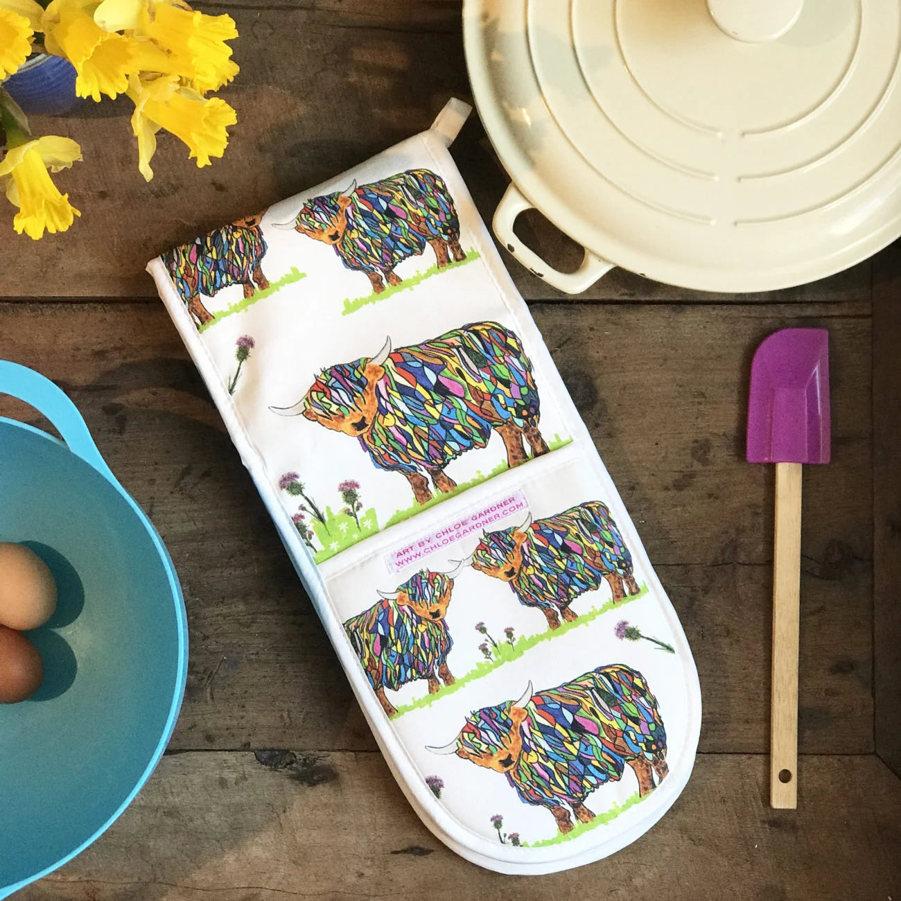 Repeat Bright Highland Cow Double Oven Glove On White on wooden background with purple and wood spatula, blue bowls with eggs in it, daffodils and cream coloured casserole dish.