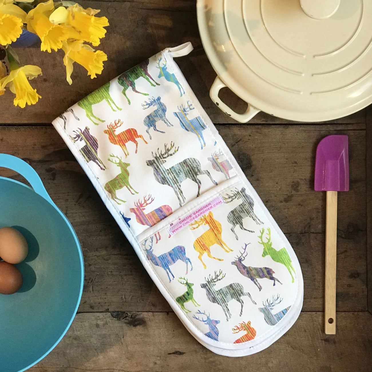 Colourful Stag Double Oven Glove (Multi) on wooden background with purple and wooden spatula. blue bowl with eggs, daffodils and cream coloured casserole dish.