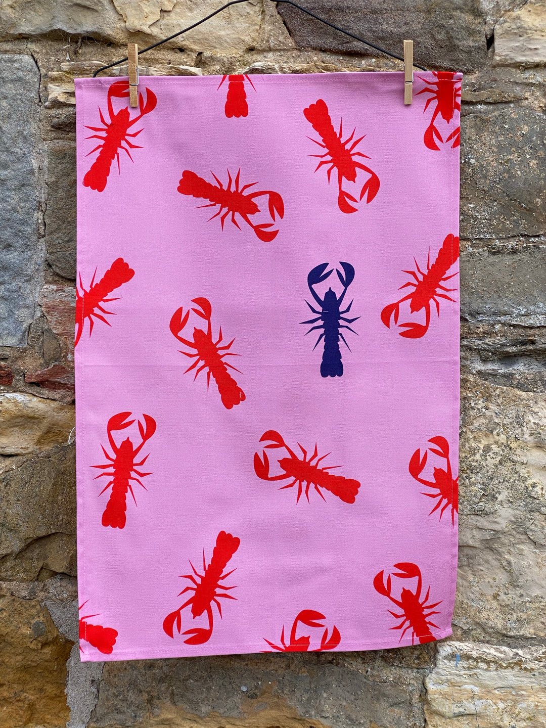 100% Cotton Pink Lobster Tea Towel hanging on pegs on stone wall background.