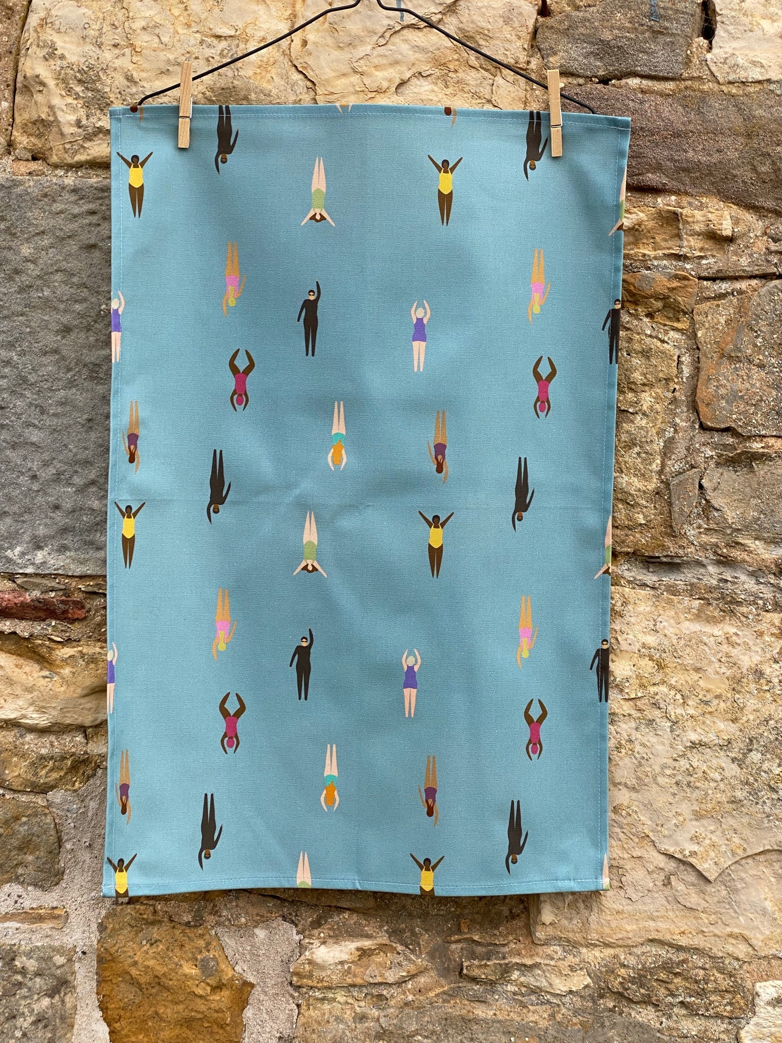 Wild Swimming Tea towel hanging by pegs on stone wall.  